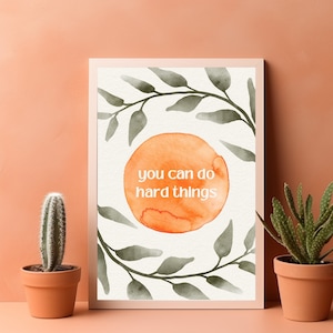 以下が含まれることがあります： オレンジ色の水彩画の円に白い文字で「you can do hard things」と書かれた額入りのプリント。緑の葉で飾られ、桃色の壁を背景に、両側に鉢植えのサボテンが置かれています。