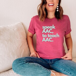 Pu&ograve; includere: Una donna indossa una maglietta rosa con il testo "speak AAC, to teach AAC." in lettere blu chiaro. &Egrave; seduta su un divano bianco con un cuscino bianco.