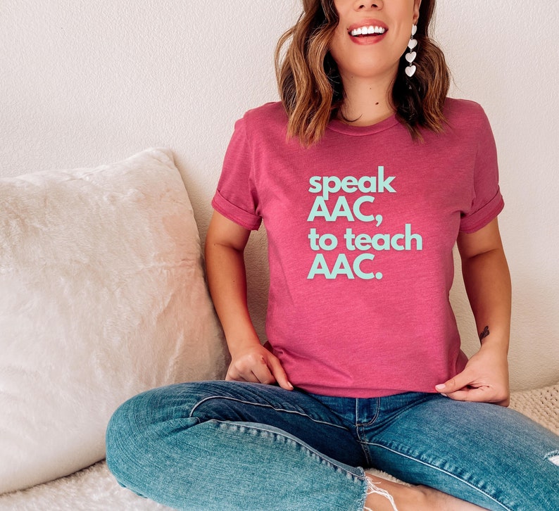 Pu&ograve; includere: Una donna indossa una maglietta rosa con il testo "speak AAC, to teach AAC." in lettere blu chiaro. &Egrave; seduta su un divano bianco con un cuscino bianco.
