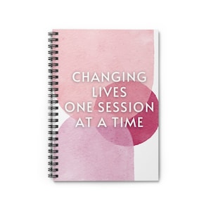 May include: A spiral-bound notebook with a watercolour design in pink and white hues. The cover displays the text "CHANGING LIVES ONE SESSION AT A TIME". The notebook has a black spiral binding.