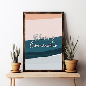 May include: A framed art print with the words "Behaviour is Communication" in white script. The artwork features abstract layers of colour: peach, teal, and grey. The print is displayed on a wooden shelf with potted plants on either side.