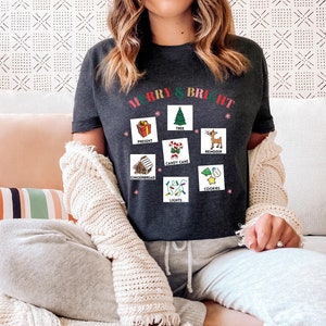 May include: Dark gray t-shirt with the words "MERRY & BRIGHT" in red and green. The shirt features Christmas-themed illustrations including a present, tree, reindeer, candy cane, gingerbread house, lights, and cookies.