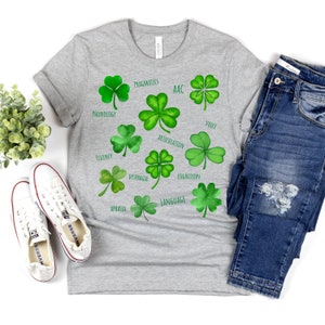 May include: A light gray t-shirt featuring watercolor illustrations of green shamrocks with text labels such as "Pragmatics" and "Articulation". The shirt is paired with blue jeans and white sneakers.