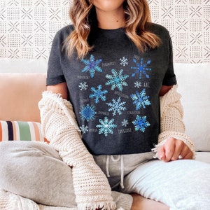 Peut inclure: Un t-shirt gris foncé avec un motif de flocon de neige. Chaque flocon de neige a un mot différent lié à l'orthophonie, comme "Articulation", "Pragmatique" et "Langage".