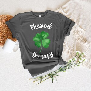 May include: Dark gray t-shirt featuring the words "Physical Therapy" in white script, with a watercolor-style green shamrock in the center. The shirt is styled with a knot at the bottom. A white mug and flowers are in the background.