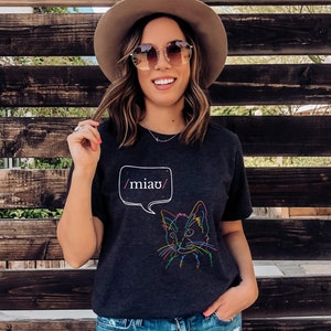 May include: A dark grey t-shirt with a colorful line drawing of a cat's face and the text "/miau/" in a speech bubble.