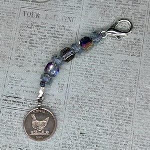 May include: A silver charm with a blue and iridescent beaded chain. The charm is a round coin with a chicken on it. The coin says "TOGOA" and "1996".