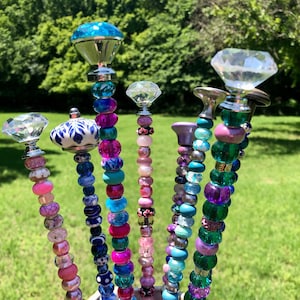 May include: A set of seven colorful garden stakes made with beads and decorative tops. The tops are made of glass and metal and feature a variety of shapes, including diamonds and round knobs. The stakes are a mix of pink, purple, blue, green, and turquoise.