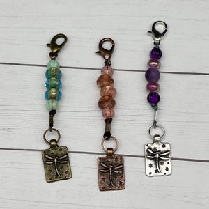 May include: Three beaded keychains with dragonfly charms. The charms are made of metal and have a rustic finish. The beads are in shades of blue, pink, and purple.