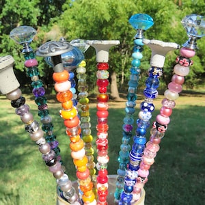 May include: A collection of colorful beaded garden stakes with various glass and metal embellishments. The stakes are arranged in a vase and feature a variety of colors, including blue, green, red, orange, and yellow.