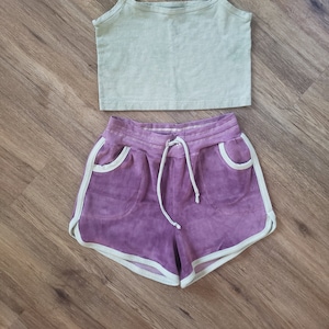 May include: A two-piece outfit with a sage green tank top and purple shorts with white trim. The shorts have a drawstring waist.