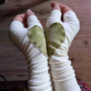 May include: A pair of off-white fingerless gloves made of a soft, stretchy fabric. Each glove features a stitched-on olive green heart-shaped patch on the back of the hand, with a thumb hole and a long, ruched sleeve that extends up the forearm.
