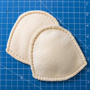 Puede incluir: Dos almohadillas de sujetador beige en forma de lágrima con bordes cosidos. Las almohadillas están hechas de una tela suave y texturizada y se colocan sobre una superficie de cuadrícula azul. Están diseñadas para soporte y forma.