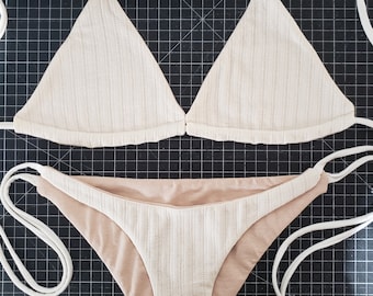 May include: A white ribbed bikini top and bottom with white straps. The bikini top is a triangle style and the bottom is a high-cut style.