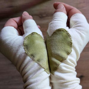 May include: A pair of off-white fingerless gloves made from a soft fabric, each featuring a stitched olive green heart-shaped patch on the back of the hand. The gloves have a thumb hole and extend to cover the wrist area with a gathered, flexible fit.