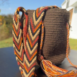 May include: A brown crocheted bag with a long, colorful strap. The strap is woven with a chevron pattern in shades of brown, red, orange, and black. The bag has a tassel made of the same yarn as the strap.