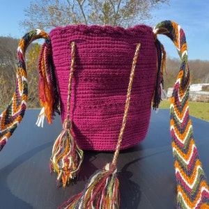 May include: A handmade, magenta crochet bag with chevron-patterned straps. The bag features colorful tassels and a scalloped top edge. The straps are a mix of white, orange, green, and navy blue.