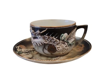 Könnte beinhalten: Ein schwarzes Tassen- und Untertassen-Set mit Drachendesign. Die Tasse hat ein detailliertes Drachenrelief mit goldenem Rand und Griff. Die Untertasse ergänzt die Tasse mit passenden Drachenmotiven und einem goldenen Rand. Ein dekoratives Teeservice.