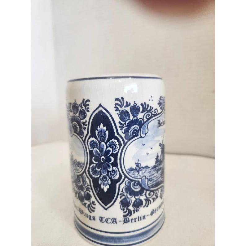 May include: A white ceramic mug with blue floral and landscape designs. The mug features a central oval design with a flower and scrollwork, and scenes of windmills and buildings. The text "Wings TCA-Berlin-Germany" is printed on the bottom.