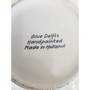 May include: Close-up of a white ceramic surface with the text "Blue Delfts Handpainted Made in Holland" in blue script. The surface has a slight curvature and a glossy finish.