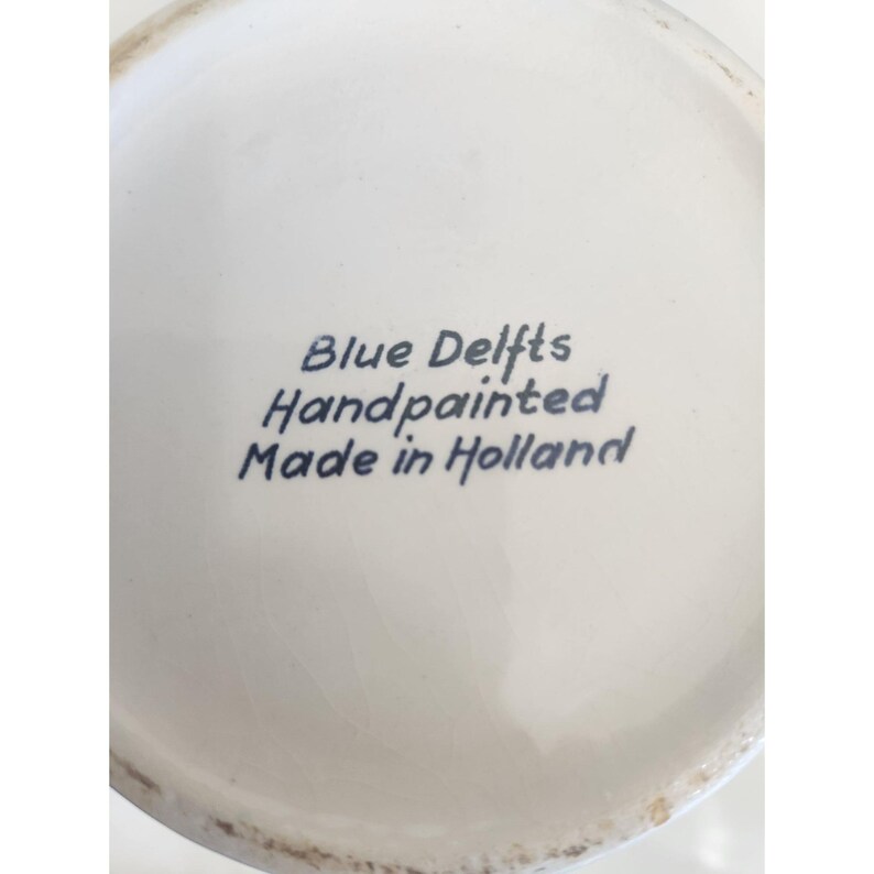 May include: Close-up of a white ceramic surface with the text "Blue Delfts Handpainted Made in Holland" in blue script. The surface has a slight curvature and a glossy finish.