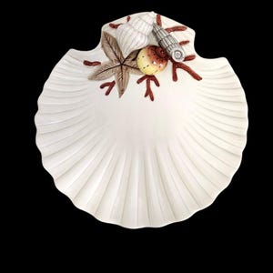 May include: A white ceramic shell-shaped dish featuring a decorative cluster of three-dimensional elements including a brown starfish, a white spiral shell, a grey textured shell, a yellow round shell, and red coral-like branches.