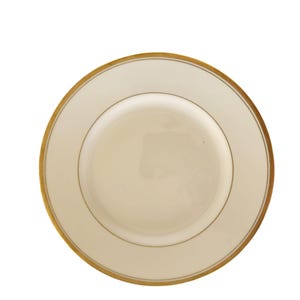 May include: A cream-coloured ceramic plate with a gold rim and a thin gold band near the edge. The plate has a simple, elegant design, suitable for formal dining. The plate is round and has a smooth surface.