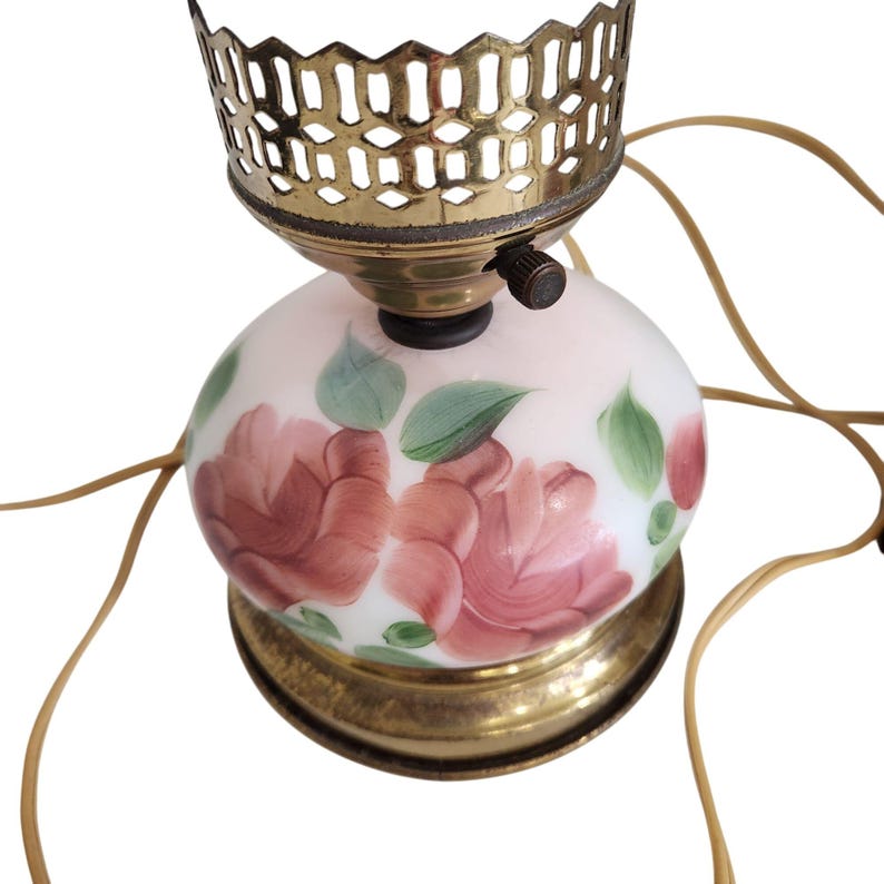 May include: A vintage table lamp with a white glass shade featuring hand-painted pink roses and green leaves. The lamp has a brass base and a brass cage around the bulb.