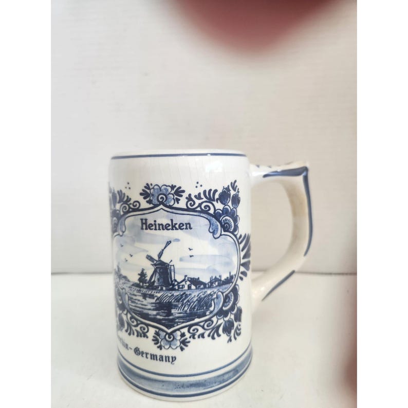 May include: White ceramic beer stein with a blue and white windmill design. The stein has a handle and the word "Heineken" is printed on the front. The bottom of the stein reads "Germany".
