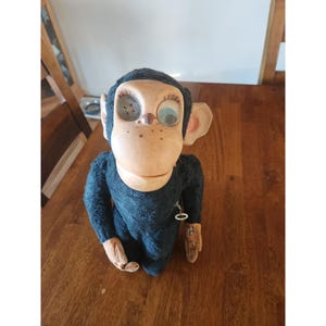 May include: A vintage wind-up monkey toy with black synthetic fur, a tan plastic face, and large ears. The monkey features one blue eye and one metallic eye, with a small metal winding key visible on its side. The hands and feet are tan plastic.