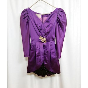 May include: A vintage purple dress with long sleeves and a V-neckline. The dress features a gold-colored brooch at the waist and a black lace skirt. The dress is hanging on a white hanger.