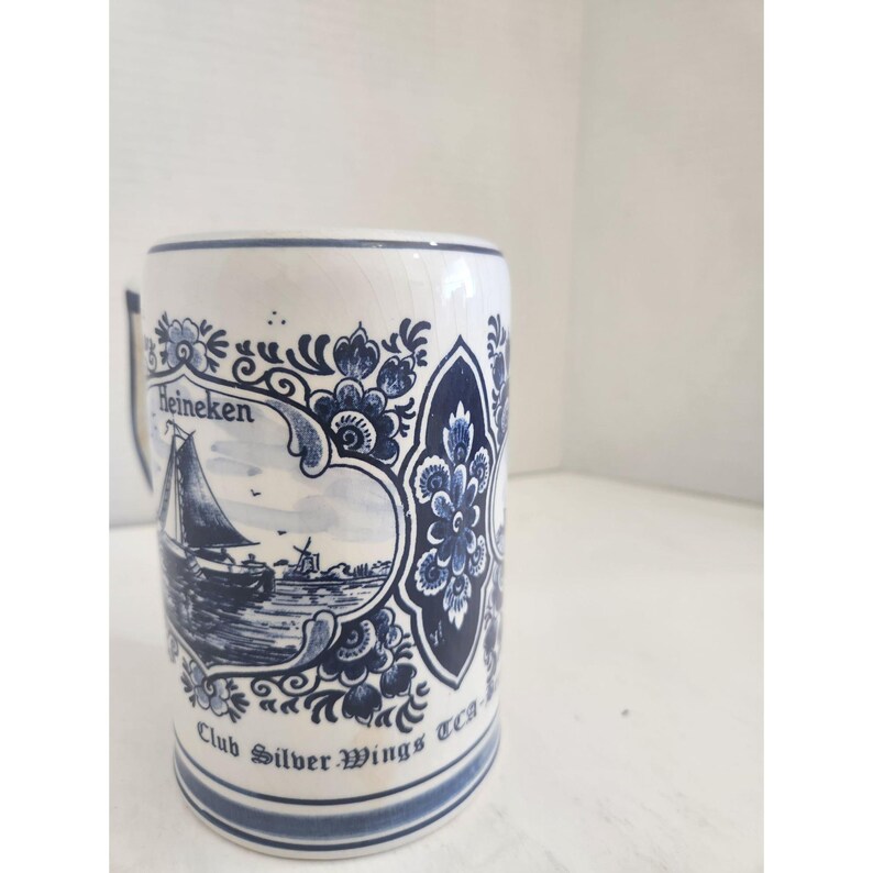 May include: White ceramic beer stein with blue floral and nautical designs. The stein features the word "Heineken" and a sailboat scene. The bottom of the stein reads "Club Silver Wings TCA".