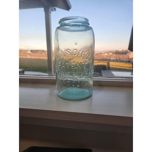 May include: A vintage, clear glass Mason's jar with a light blue tint. The jar has embossed text that reads "MASON'S PATENT NOV. 30TH 1858". The jar is cylindrical with a wide mouth and sits on a white windowsill.
