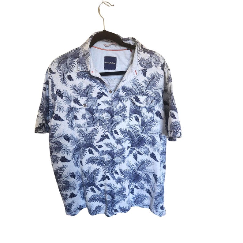 K&ouml;nnte beinhalten: Ein kurz&auml;rmeliges Poloshirt mit wei&szlig;er Grundfarbe und einem dunkelblauen Muster aus Palmbl&auml;ttern und Tannenzapfen. Das Shirt hat einen Kragen, eine kurze Knopfleiste und zwei Brusttaschen mit Klappen. Das Tommy Bahama Etikett ist am Hals sichtbar.