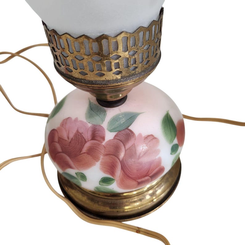 May include: A vintage table lamp with a white frosted glass shade and a hand-painted floral design. The base is a white glass globe with pink roses and green leaves. The lamp has a brass base and a brass shade holder.