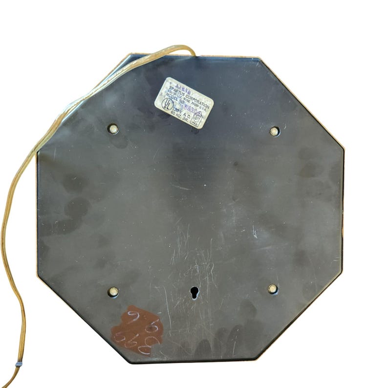 K&ouml;nnte beinhalten: Ein achteckiges Objekt aus schwarzem Metall mit goldfarbener Umrandung. Auf der Oberseite ist ein Aufkleber mit Text angebracht. Ein Netzkabel erstreckt sich von der oberen linken Ecke. Die Unterseite hat ein Schl&uuml;sselloch und mehrere Schraubenl&ouml;cher. Zahlen sind mit brauner Tinte geschrieben.