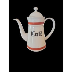 May include: White ceramic coffee pot with a lid and handle. The pot has a red band around the top and bottom, with the word "Cafe" in black script on the front. The spout is long and curved.