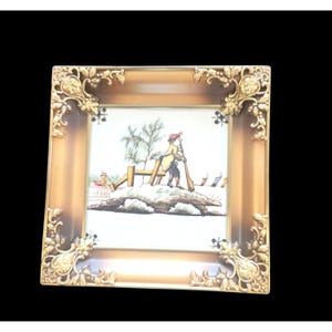 May include: A framed print featuring a winter scene with a figure shoveling snow. The frame is a warm brown with ornate gold corner accents. The artwork depicts a person in a yellow coat and red hat, with a fence and snow-covered ground.