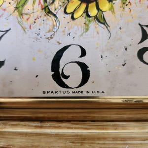 K&ouml;nnte beinhalten: Eine Vintage-Metallschale mit einem bemalten Blumendesign und der schwarzen Zahl 6. Die Schale hat einen goldfarbenen Rahmen und den Text "SPARTUS MADE IN U.S.A."