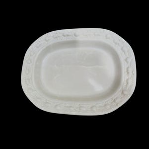 Op de afbeelding: Een witte ovale serveerschaal met een gedetailleerde, verhoogde rand. De rand heeft een herhalend patroon van kleine, getextureerde ontwerpen. Het oppervlak van de schaal is glad en reflecterend, met een subtiel, reliëf ontwerp in het midden.