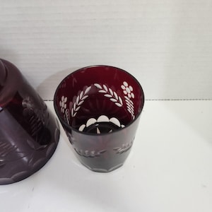 May include: A deep red glass with white floral etched patterns. The glass has a hexagonal base and a cylindrical shape. The etched design includes leaves and flower-like patterns. The glass is part of a set, with a matching piece visible on the left.