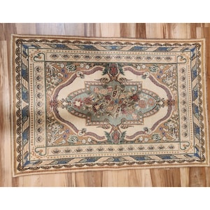 May include: Rectangular beige rug with an ornate floral design. The rug features a central floral medallion in shades of green, red, and blue, surrounded by a cream-colored border and detailed with intricate patterns and a scalloped edge.
