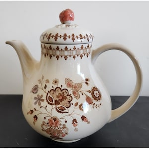 Puede incluir: Una tetera de cerámica blanca con un patrón floral en marrón y rosa. La tetera tiene una tapa con un pequeño pomo rosa en la parte superior.