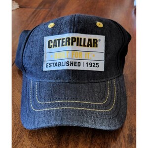 Peut inclure: Une casquette de baseball en denim noir avec des coutures jaunes et un patch blanc qui indique "Caterpillar® Built For It. Established | 1925".