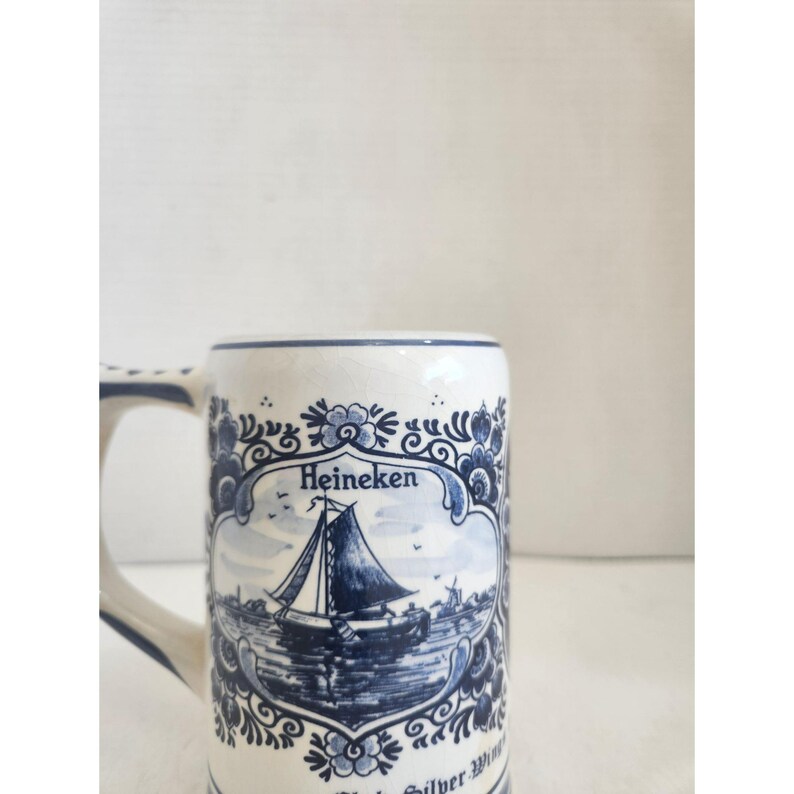 May include: White ceramic beer stein with a blue and white nautical design. The stein features a sailboat scene, floral accents, and the word "Heineken". The handle and rim are also trimmed in blue.