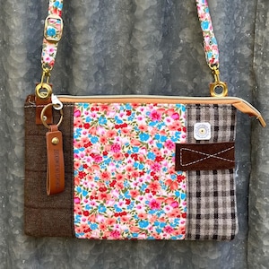 May include: A patchwork crossbody bag with a floral strap and body. The bag features brown tweed, floral, and plaid fabric panels. A leather tag with text hangs from the zipper. The bag has gold-tone hardware.
