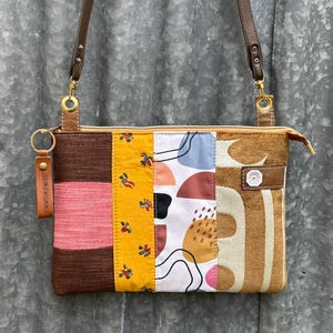 May include: A rectangular crossbody bag with a patchwork design. The bag features a variety of fabrics in brown, pink, yellow, and patterned sections. It has a brown leather strap and a zipper closure. A small leather tag hangs from the zipper.