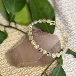 May include: A delicate bracelet featuring small, translucent, pale yellow faceted beads with dark inclusions, linked together by thin gold-toned wire. The circular jewelry piece includes a simple hook-and-eye clasp mechanism for closure.