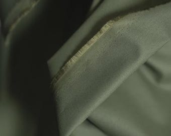 May include: Close-up of olive green fabric with a textured surface and frayed edges. The fabric is folded, creating shadows and highlighting the material's texture. The image showcases the color and detail of the textile.