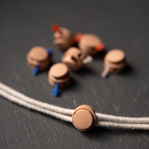 May include: A close-up of a beige cord toggle, with a white cord running through it. The toggle is on a dark gray surface, with several other beige toggles scattered around it.